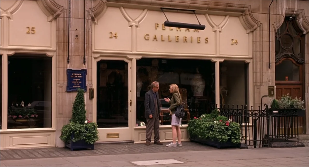 Scoop (2024) movie still: medium — An elegant London storefront with cream-colored masonry and gold-lettered signage stands on a sidewalk. An older…