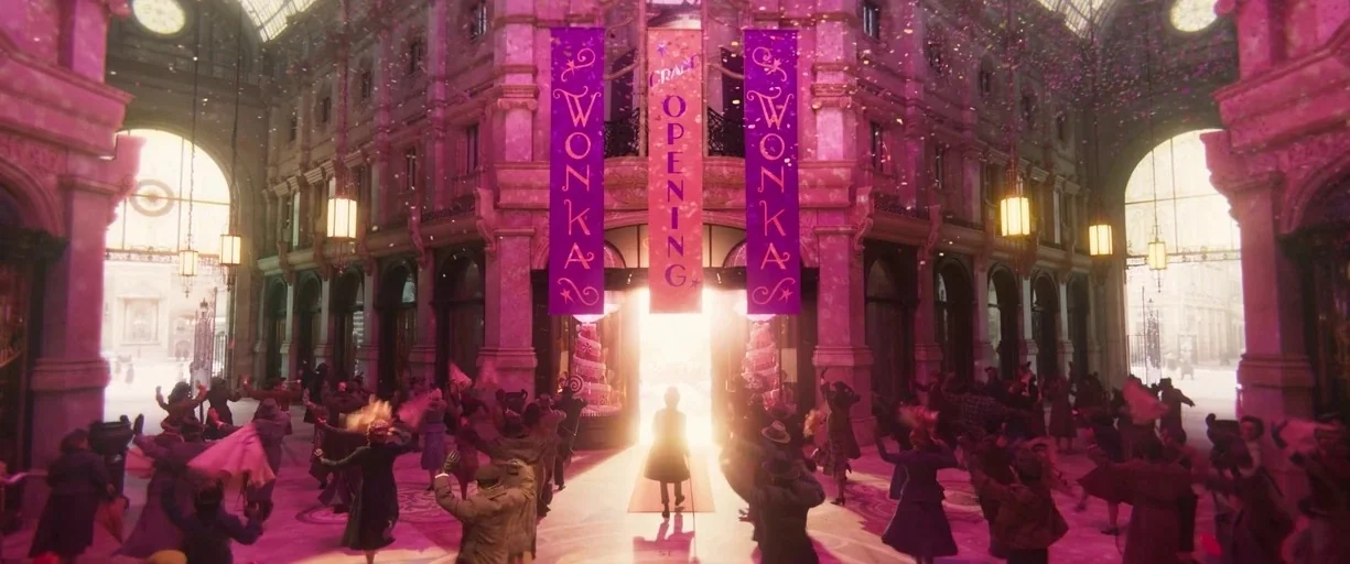 Wonka (2023) movie still: wide — An ornate, high-ceilinged shopping arcade is filled with a cheering crowd under confetti. A man in a dark coat…