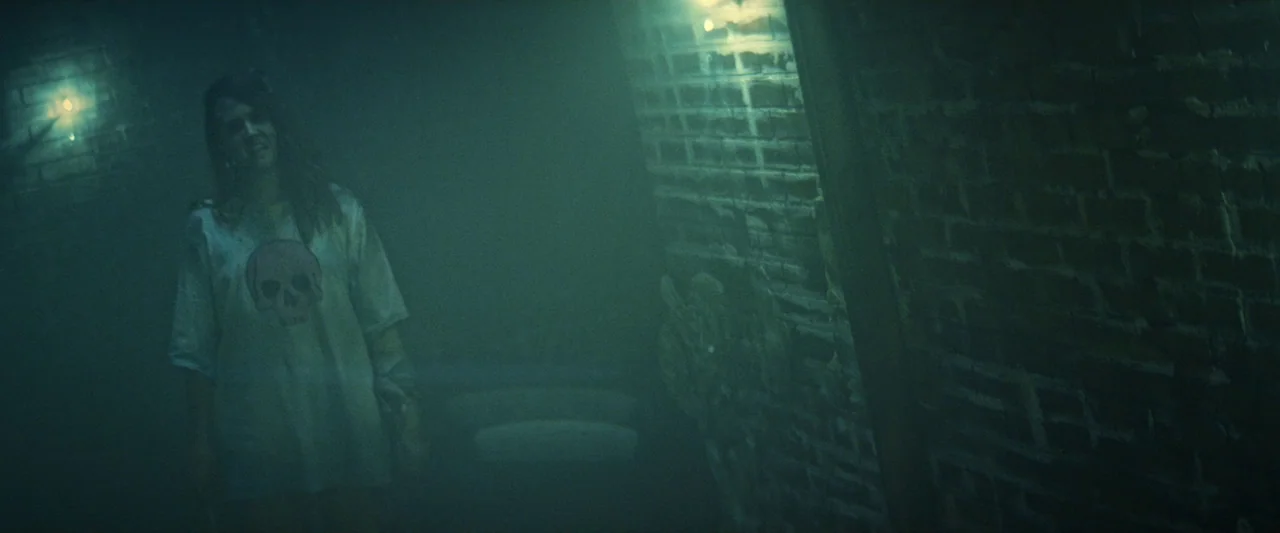 Revealer (2022) movie still: medium — A dark, atmospheric basement space is filled with dense haze and low green light. A young woman stands in the…