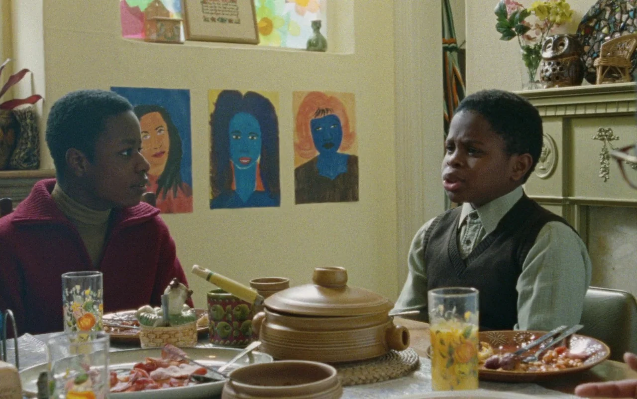 Education (2020) movie still: medium — A residential room features light-colored walls decorated with colorful child-created portrait paintings. Two…