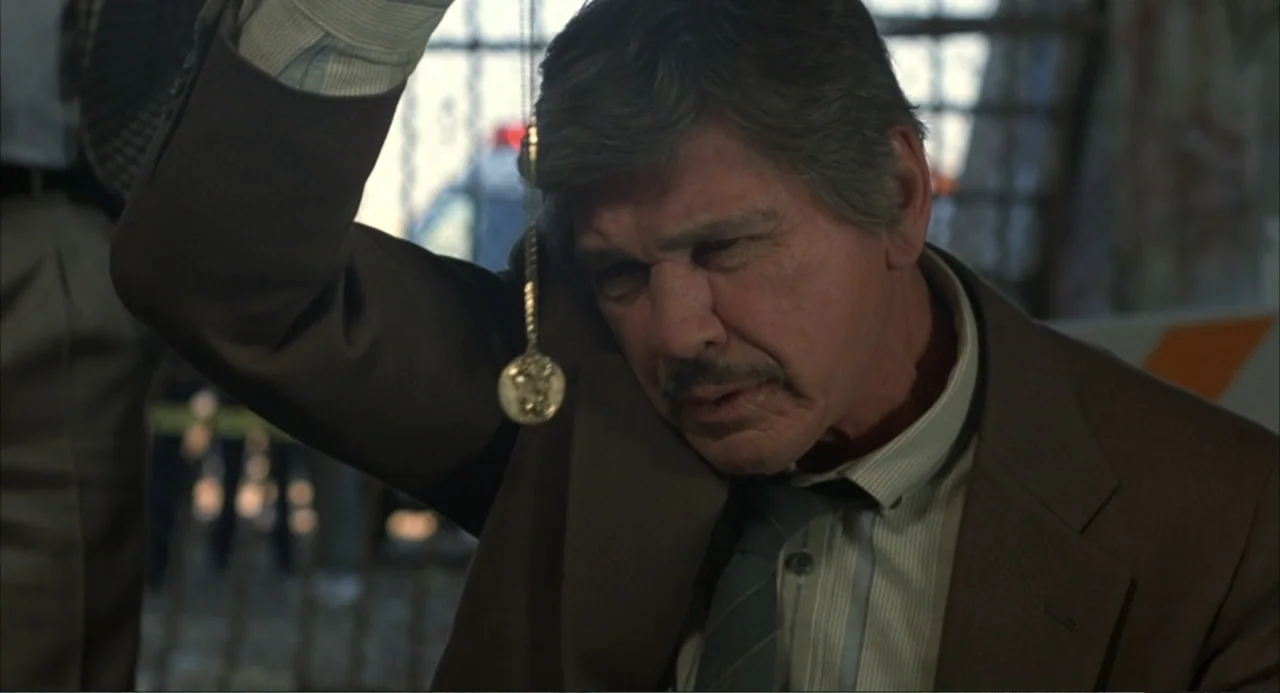 Murphy's Law (2009) movie still: medium — An older man wearing a brown suit stands indoors near a window. He holds a small circular gold medallion…