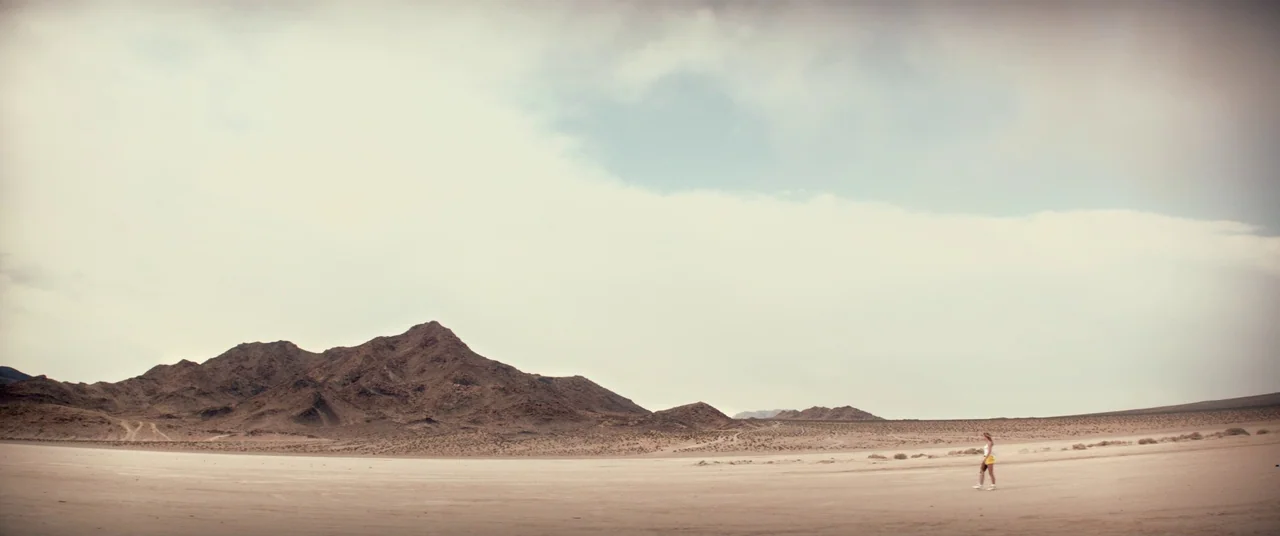 The Bad Batch (2016) movie still: wide — A vast, flat desert basin with rugged, barren mountains rising in the distance. A lone person walks slowly…