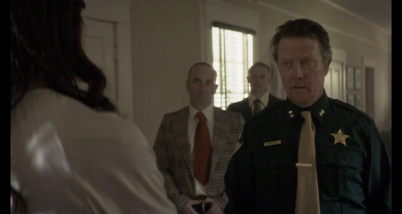 The Poison Rose (2019) movie still: medium — A dimly lit office features vintage furniture and a window casting muted daylight. A police officer in…