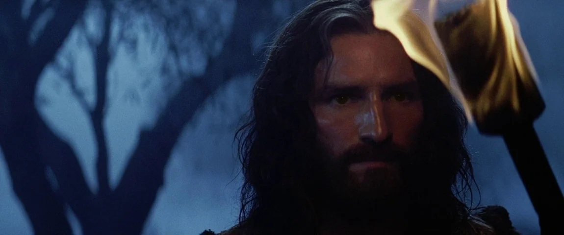 The Passion of the Christ (2004) movie still: closeup — A man with dark hair stands outdoors in the cool blue light of night. He holds a lit torch…