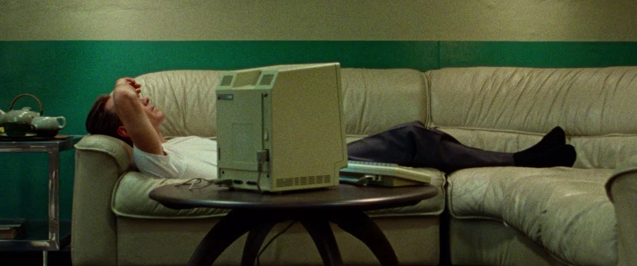 Steve Jobs (2015) movie still: medium — A living room features a green wall and a white leather sectional sofa. A man rests on the sofa while an old…