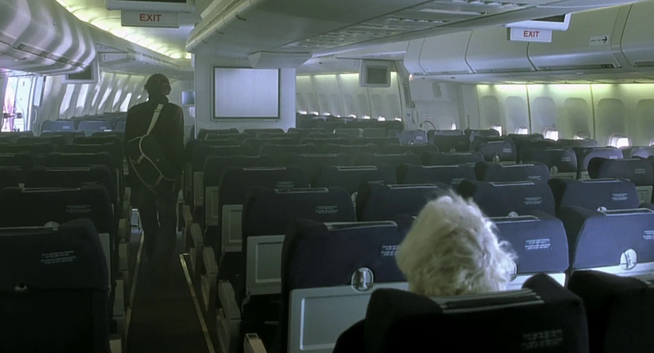 The Diving Bell and the Butterfly (2007) movie still: wide — The interior of an airplane cabin shows rows of blue seats and exit signs. A man walks…