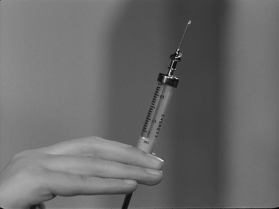 Brink of Life (1958) movie still: closeup — A medical setting with a clinical grey background. A hand holds a glass syringe with graduation markings.