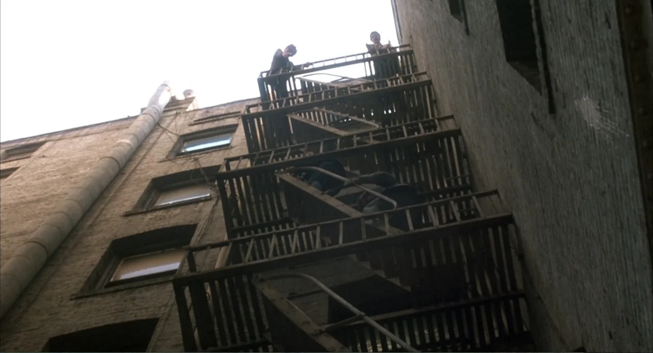Murphy's Law (2009) movie still: wide — A tall brick apartment building exterior features a dark metal fire escape attached to it. Three individuals…