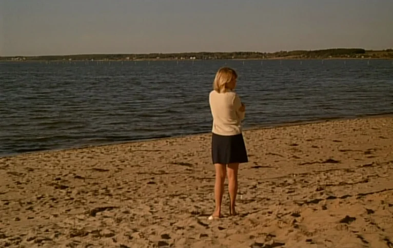 Giliap (1975) movie still: fullbody — A wide beach at the edge of a large body of water during sunset. A lone blonde woman in a skirt and sweater…