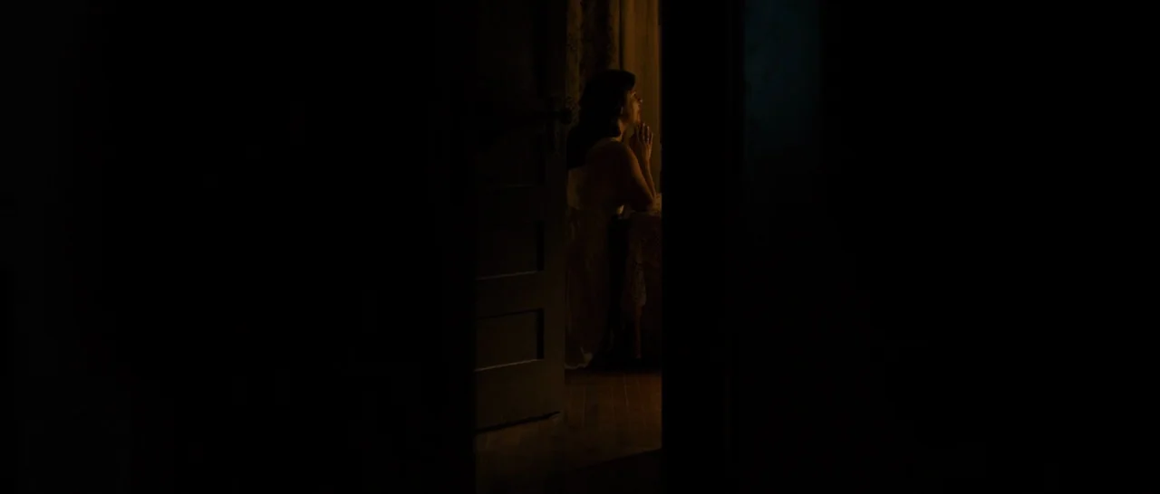 Unbroken (2014) movie still: wide — A woman is positioned in the light of an open doorway within a dark space. She kneels or sits on the edge of a…