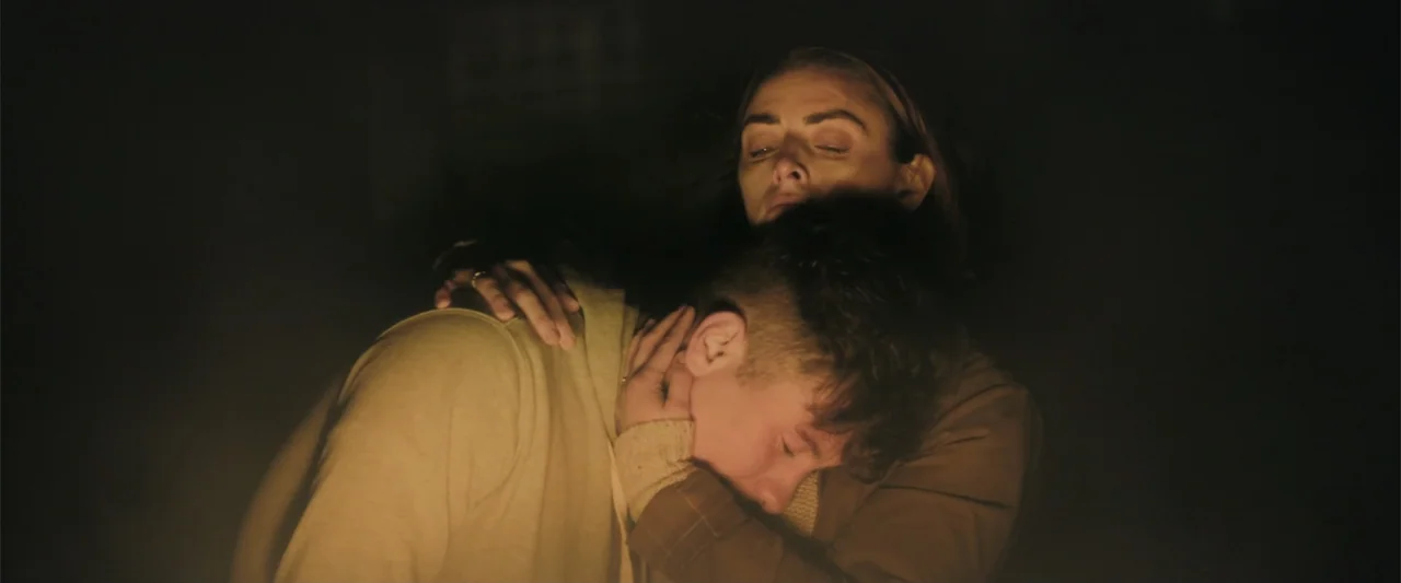 Bring Them Down (2025) movie still: medium — A dark outdoor space illuminated by a warm fire source at night. A woman embraces a young man who rests…