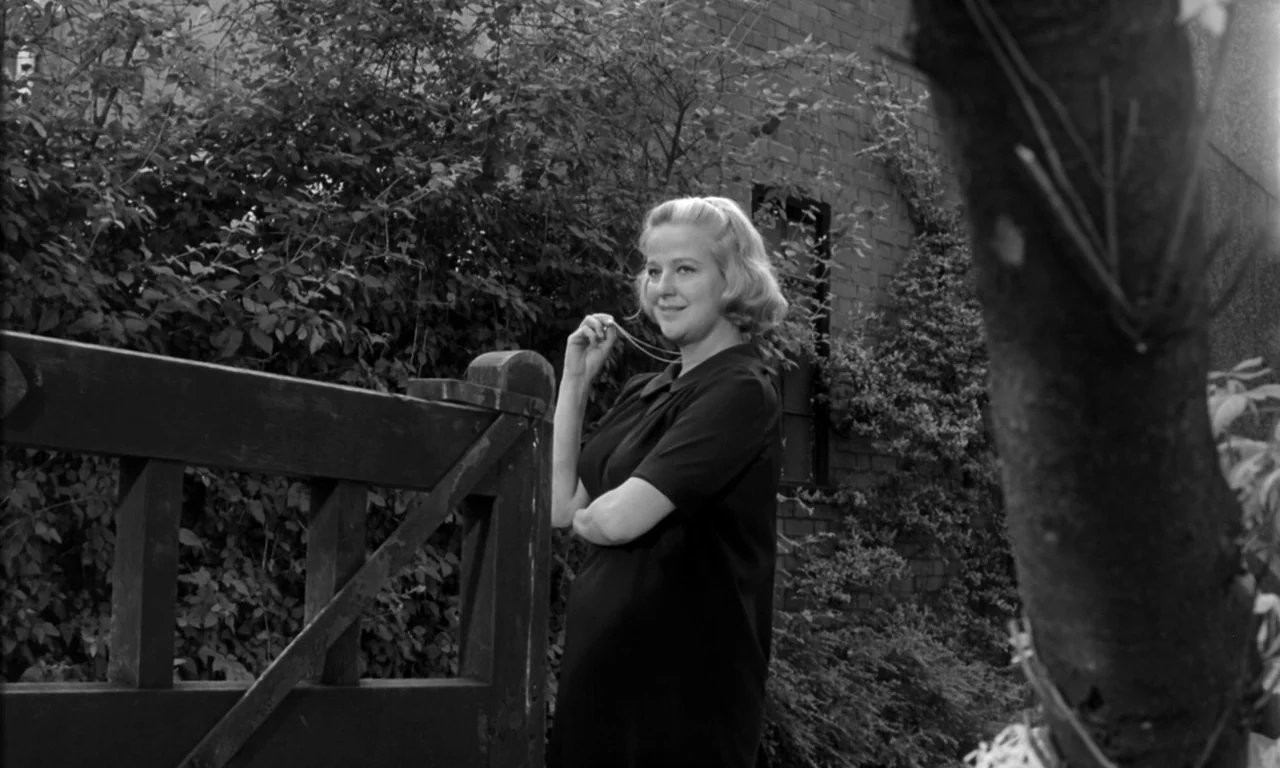 Seance on a Wet Afternoon (1964) movie still: medium — A woman stands in a lush yard with dense foliage and a brick wall. She holds a delicate chain…