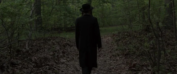 To Dust - Nightmares (2018) movie still: fullbody — A dense green woodland trail covered in dry leaves spans the frame horizontally. A man wearing a…