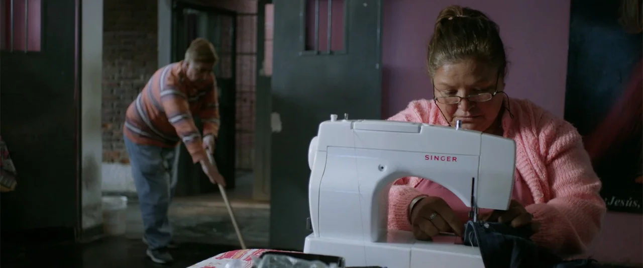 I Trust in You (2016) movie still: medium — A utilitarian room features a white sewing machine on a table in the foreground. A woman operates the…