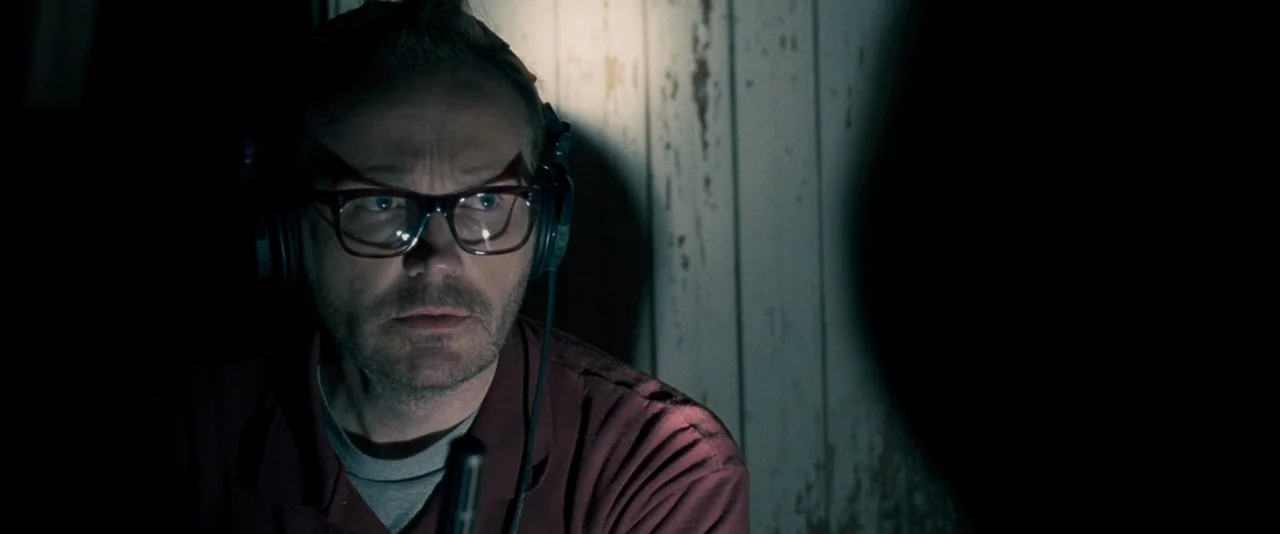 The Innkeepers (2011) movie still: closeup — A man sits in a dimly lit space against a white paneled wall. He wears professional audio headphones…