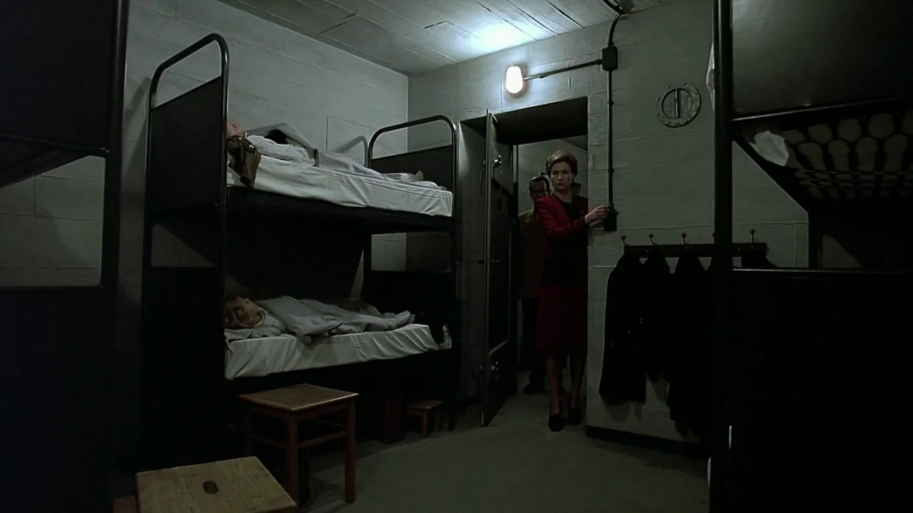Downfall (2004) movie still: wide — A austere concrete room contains metal bunk beds with occupants visible under white linens. A woman in a red…