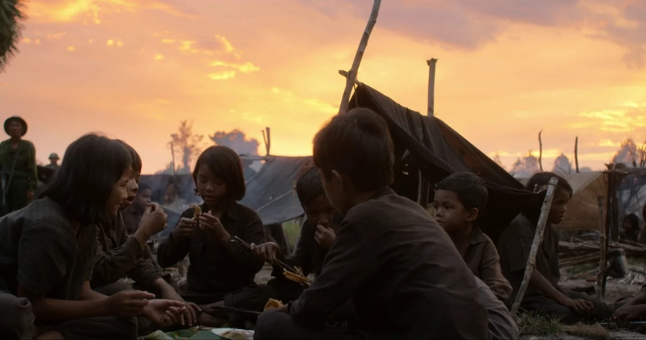 First They Killed My Father (2017) movie still: medium — A group of children sits in a makeshift camp at sunset among crude shelters. They are…