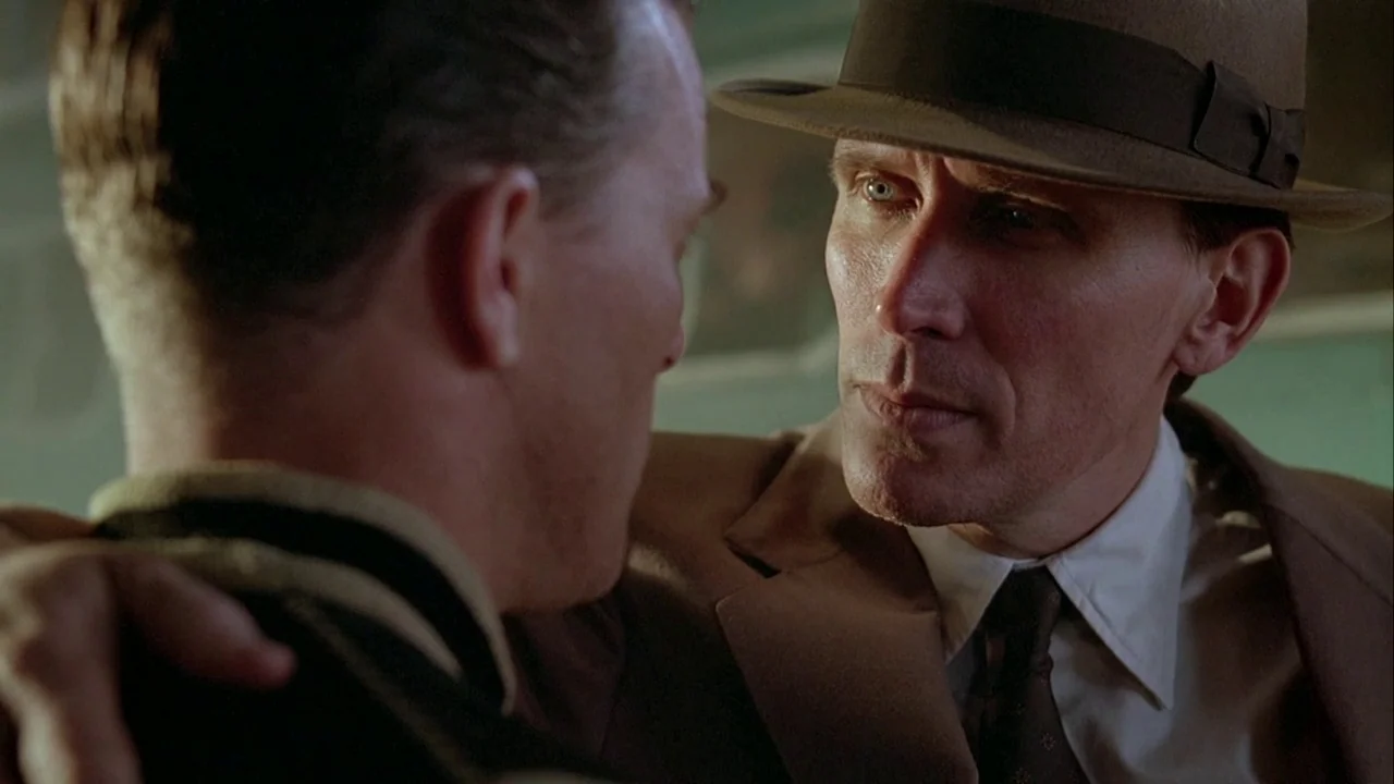 Naked Lunch (1991) movie still: medium — A dimly lit interior space with muted green walls showing worn surfaces. A man in a brown fedora leans…