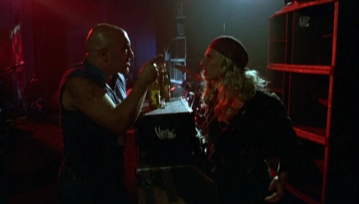 Highway 61 (1991) movie still: medium — A dark music club features music equipment, racks, and red lighting. Two men confront one another over a…