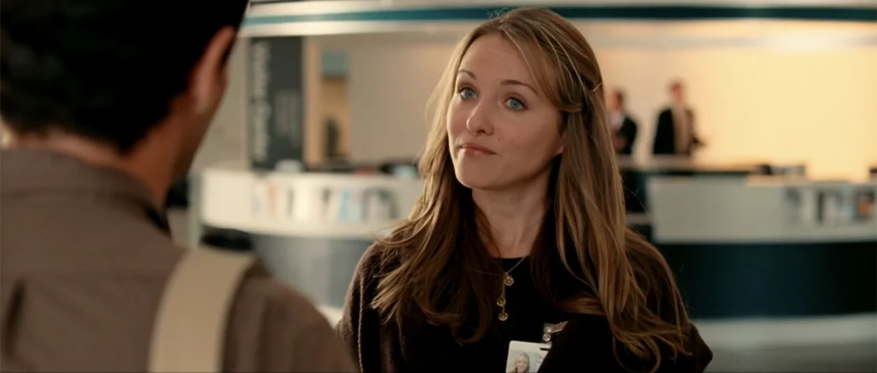 The Rebound (2009) movie still: medium — A brightly lit hospital or office reception area with a white curved desk. A woman with long brown hair…