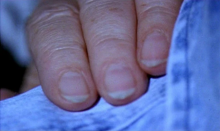 Jacquot de Nantes (1991) movie still: closeup — Three fingers of a human hand rest upon the surface of textured blue fabric. The focus is sharp on…