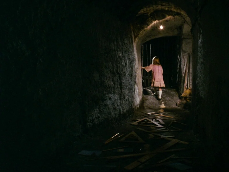 Alice (1988) movie still: wide — A dark subterranean tunnel made of rough, uneven stone walls and a dirt floor. A young girl walks away through an…