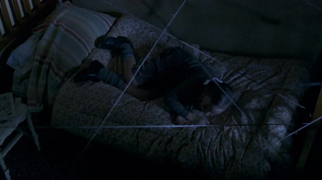 Spider (2002) movie still: medium — A child's bed features a patterned comforter and a striped pillow in a dark space. The child rests on the…