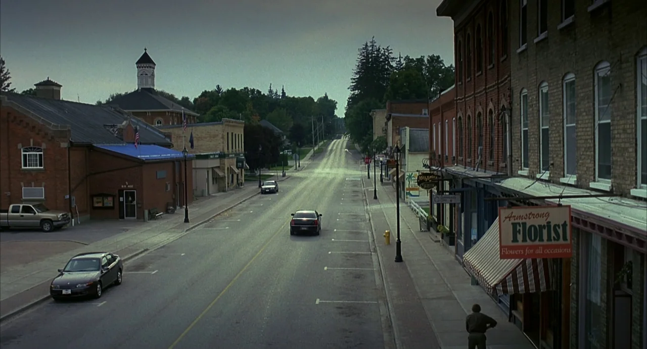 A History of Violence (2005) movie still: establishing — A quiet small-town street features brick buildings with storefronts on an overcast day. A…