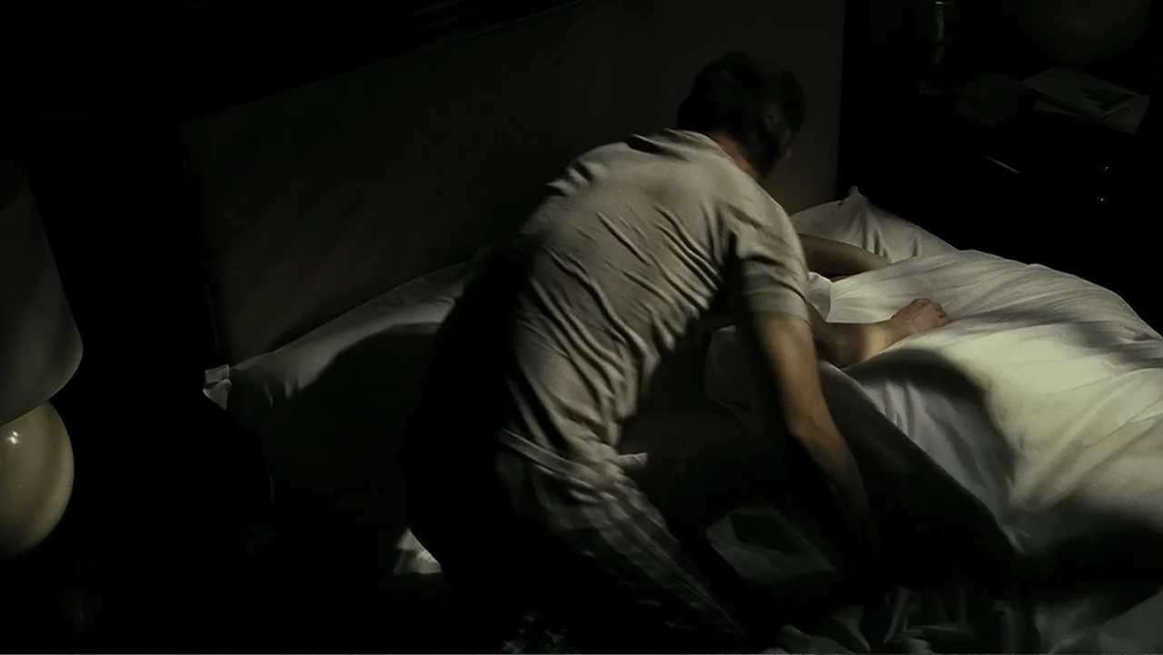 Mr. Brooks (2007) movie still: medium — A dimly lit bedroom features a bed with white linens and a dark headboard. A man in a t-shirt kneels on the…