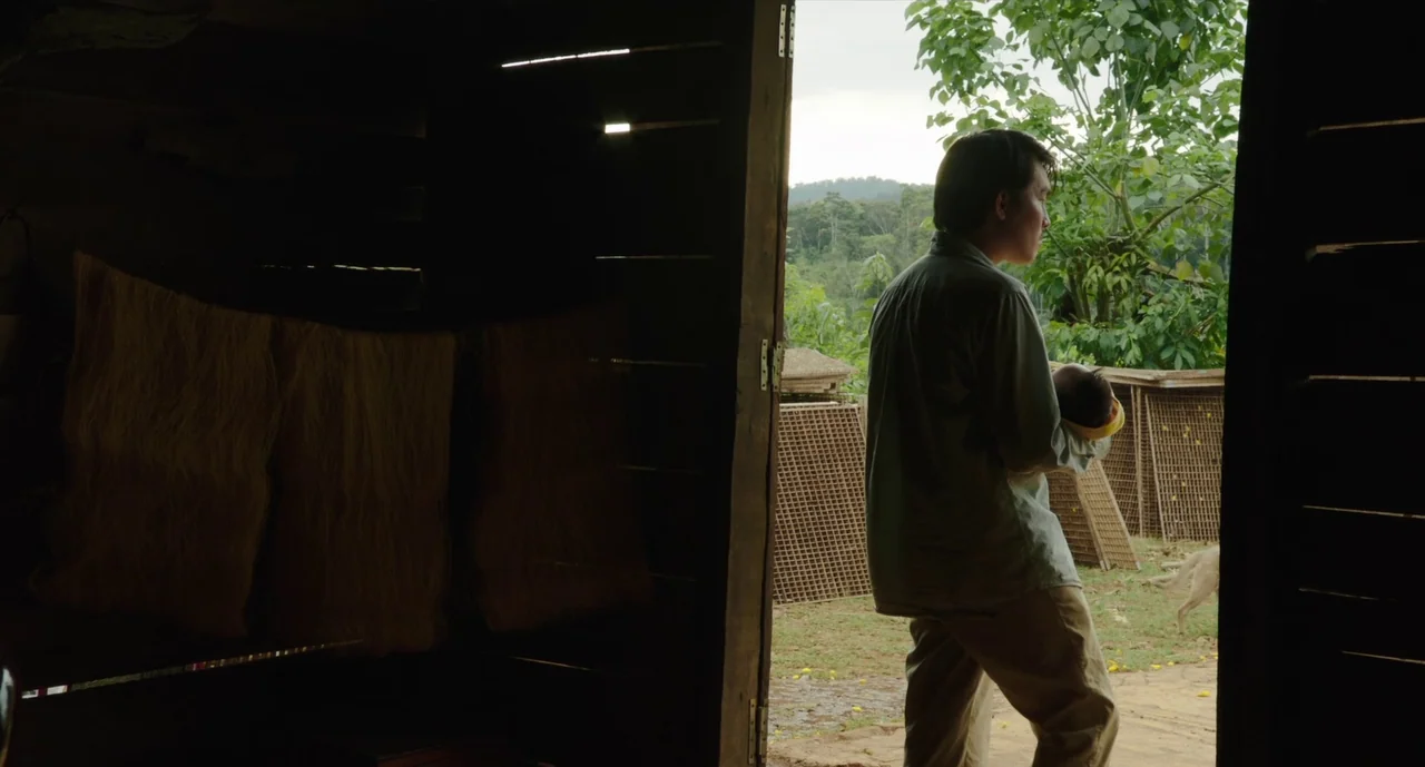 Inside The Yellow Cocoon Shell (2023) movie still: fullbody — A dark wooden structure opens onto a rural area with lush greenery. A man holds a…