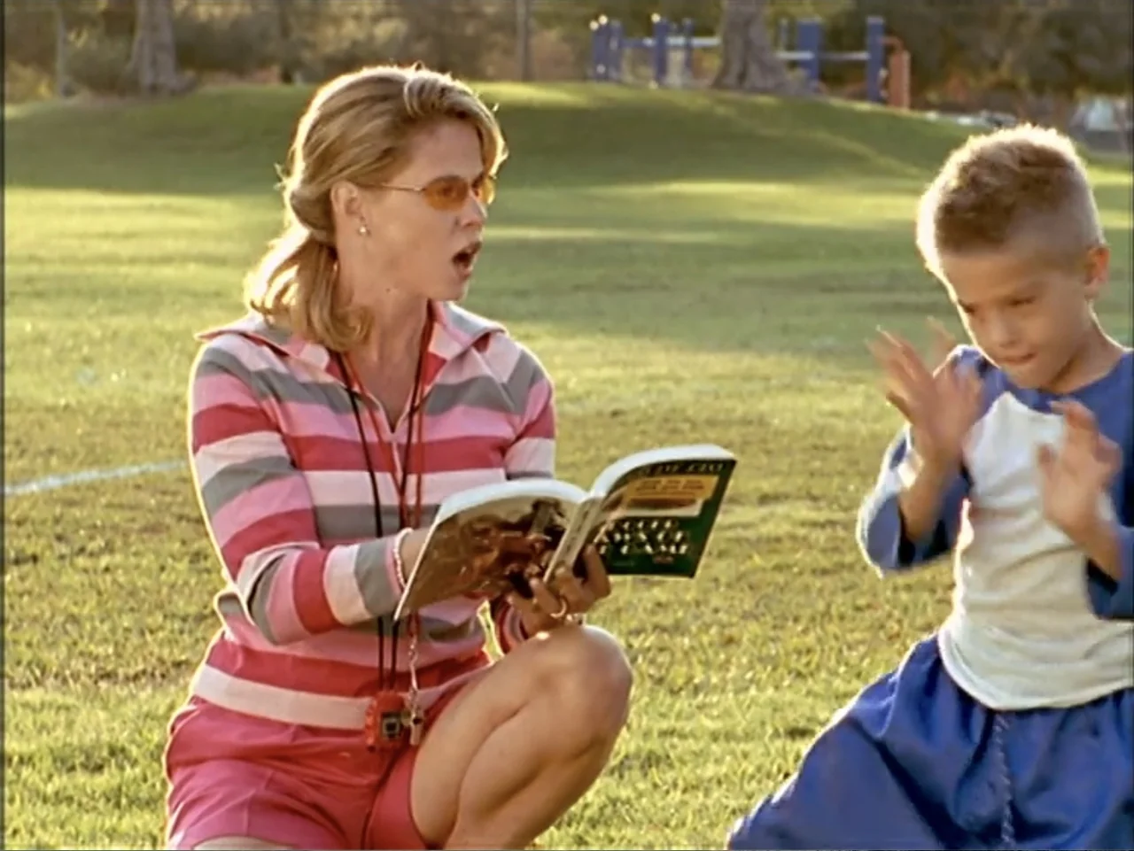 Just 4 Kicks (2003) movie still: medium — A grassy field in a park during a sunny afternoon. A woman kneels on the grass reading a book to a young…