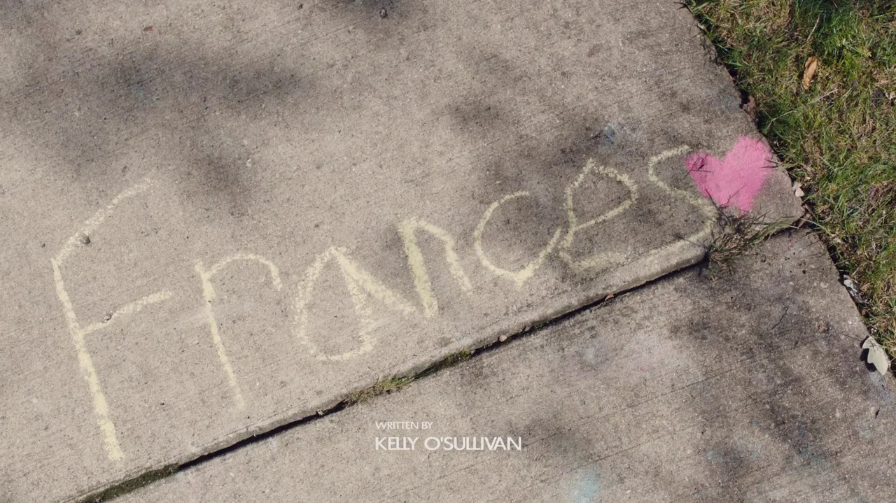 Saint Frances (2019) movie still: closeup — A textured concrete sidewalk surface shows yellow chalk text and a pink heart drawing. The word Frances…