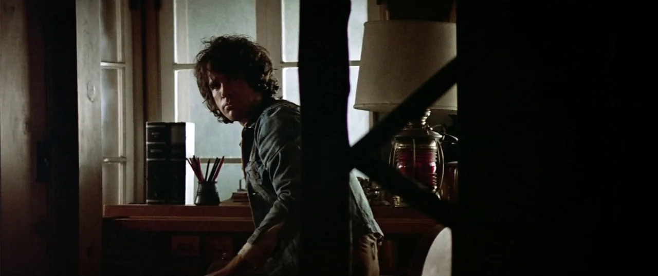 The Parallax View (1974) movie still: medium — A dimly lit room features a wooden desk placed directly in front of windows. A man in a denim jacket…