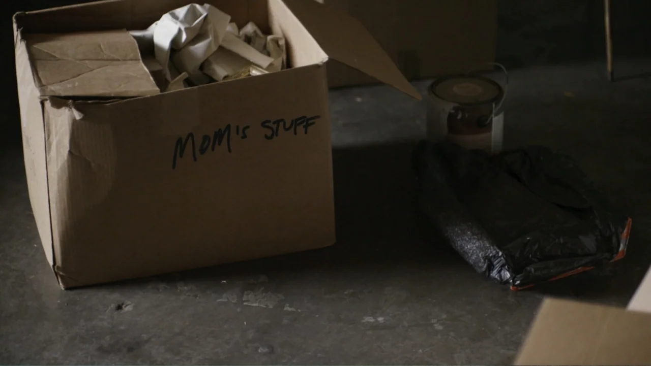 Totem (2022) movie still: medium — A brown cardboard box with handwritten text sits on a dark concrete floor. Next to the box is a paint can and a…