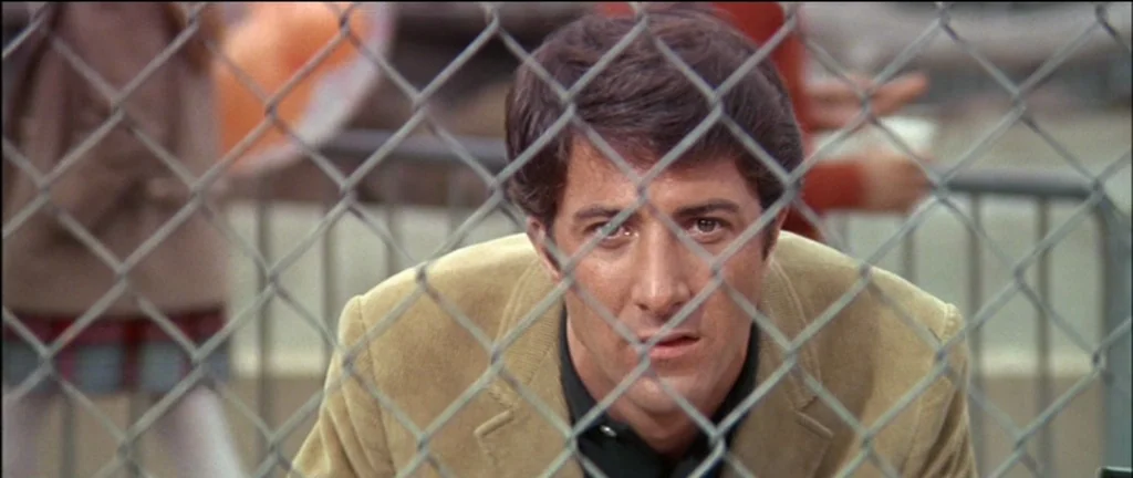 The Graduate (1967) movie still: closeup — A man stands behind a metal chain-link fence outdoors. The subject stares directly through the grid…