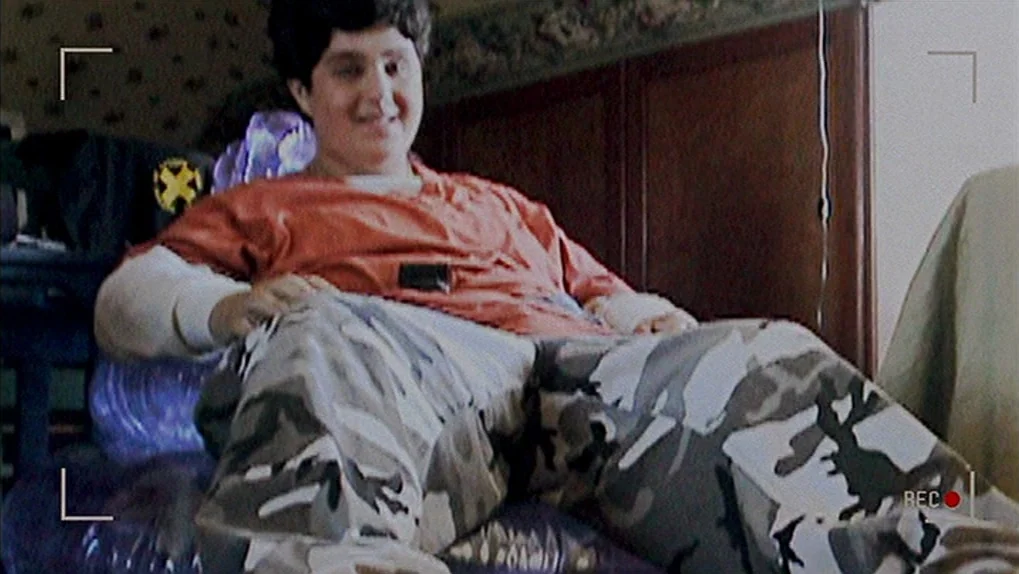 Mean Creek (2004) movie still: medium — A person sits on a purple inflatable chair in a room with floral wallpaper. The person wearing a t-shirt and…
