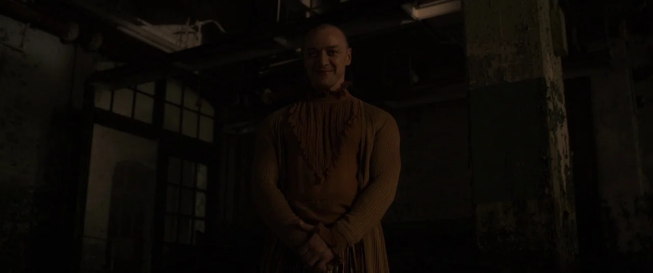 Glass (2019) movie still: medium — A dimly lit basement room with concrete pillars and dark industrial shadows. A man with a shaved head stands…