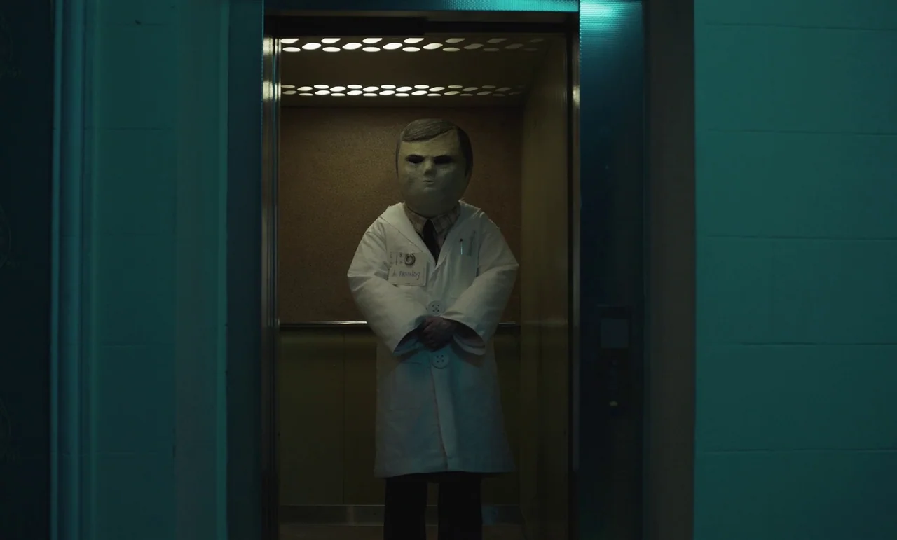 The Silent Twins (2022) movie still: fullbody — A sterile industrial elevator features plain walls and a recessed ceiling lighting panel. A person…