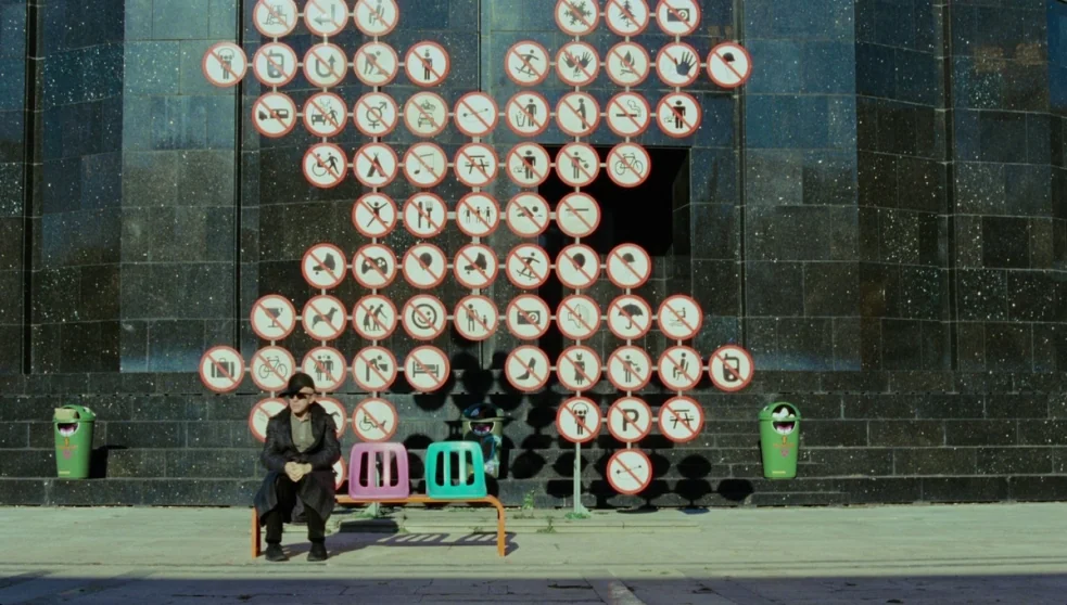 The Zero Theorem (2013) movie still: wide — A dark granite wall is covered in numerous circular red-bordered prohibition signs. A man sits alone on…