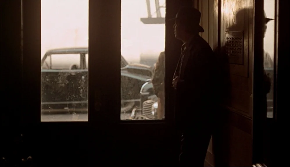 The Godfather (1972) movie still: medium — An interior entrance with wood paneling faces outward toward a street through windows. A man stands in…