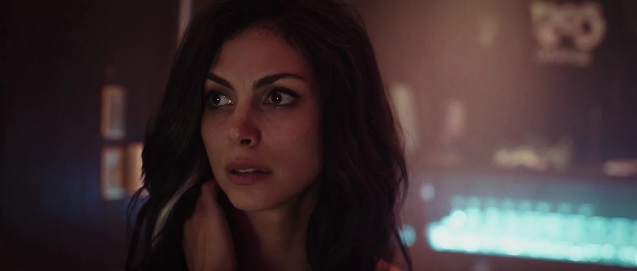 Deadpool (2016) movie still: closeup — A dimly lit nightlife venue features dark walls and blurred cyan neon signage. A woman with dark hair stands…