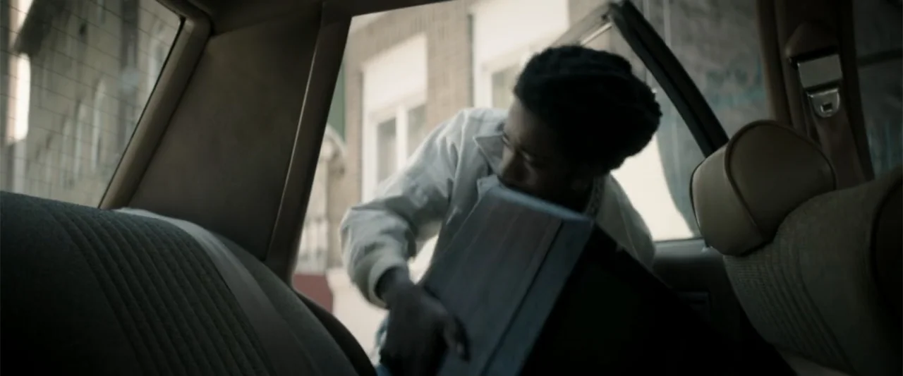 Crown Heights (2017) movie still: medium — The interior of a car back seat shows an open door to a sidewalk. A young man reaches into the space to…