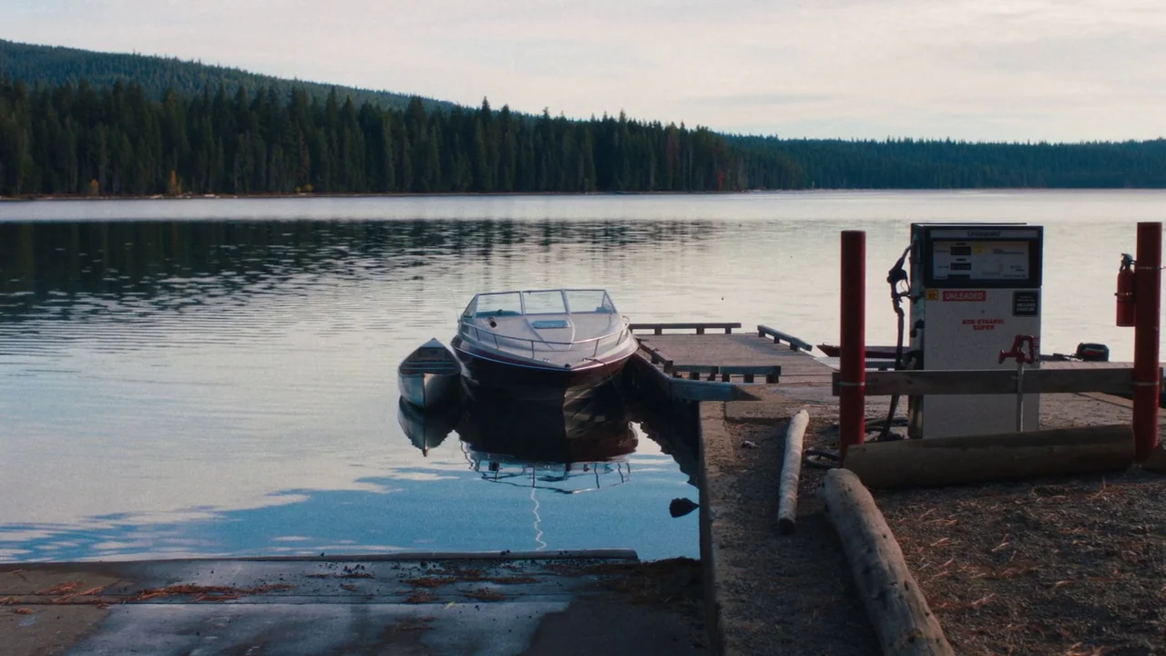 Night Moves (2013) movie still: wide — A boat dock extends into a calm, wide lake bordered by dense pine forests. A fuel pump station stands at the…