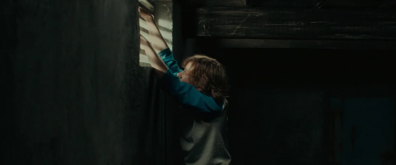 The Black Phone (2021) movie still: medium — A dark subterranean concrete space features a high rectangular window with metal slats. A young boy in…