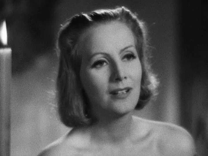 Queen Christina (1933) movie still: closeup — A woman stands in a darkened room illuminated primarily by a nearby flickering candle. She looks…