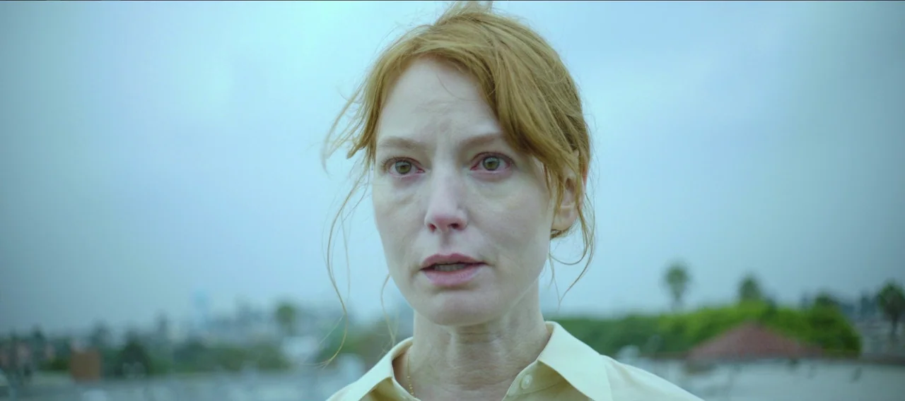 Fuzzy Head (2023) movie still: closeup — An outdoor space with blurred vegetation and buildings in the distant, overcast background. A woman with…