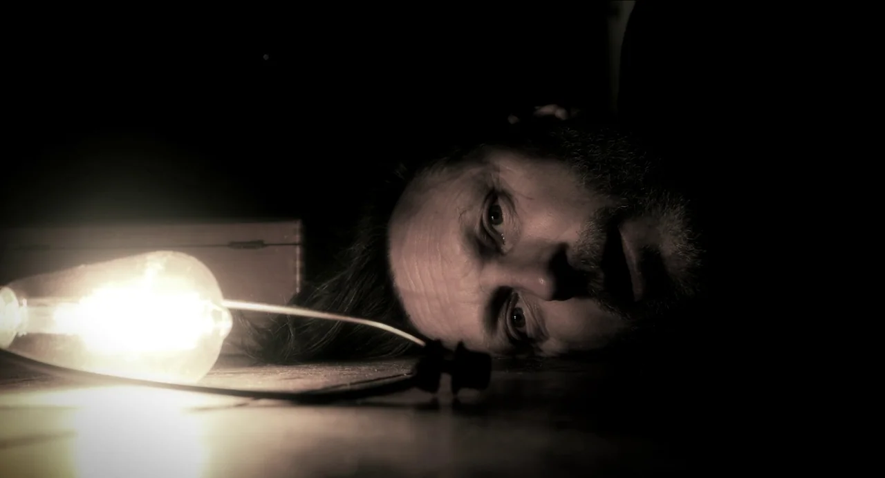 Where The Devil Roams (2023) movie still: closeup — A man lies prone on a dark floor in an otherwise unlit space. A single incandescent lightbulb…