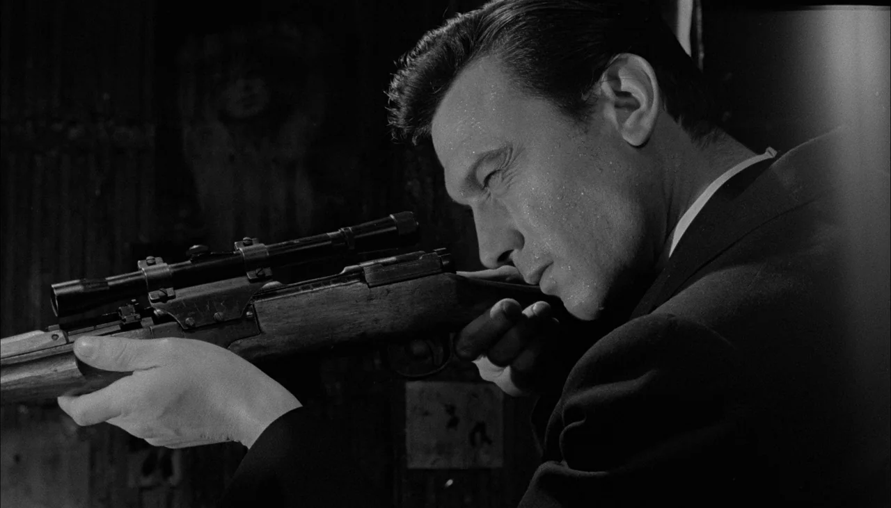 The Manchurian Candidate (1962) movie still: medium — A man is positioned in a dark room holding a sniper rifle. He aims the weapon toward the right…