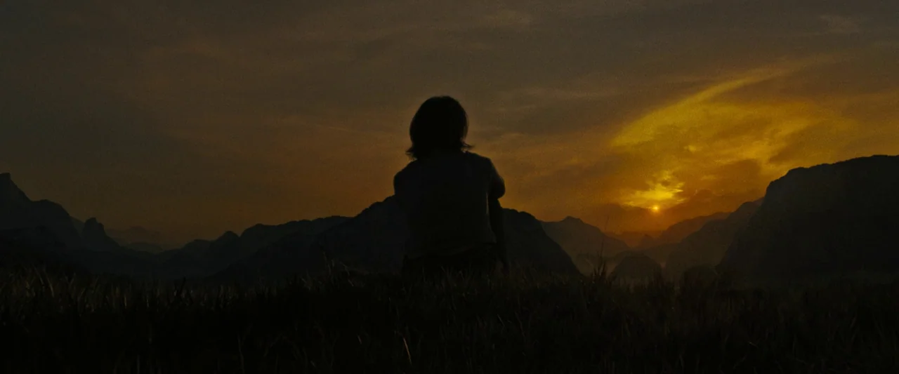 Alien: Romulus (2024) movie still: wide — A grassy field with a distant mountain range sits beneath a dark sunset sky. A solitary figure sits on the…