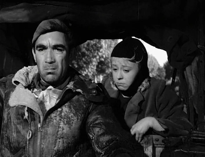 La Strada (1954) movie still: medium — Two people are positioned inside the open cargo space of a military vehicle. They look forward while seated…